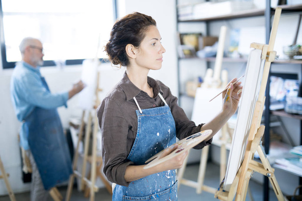 Nuori vakava nainen keskittyy maalaamaan kuvan maalaustelineeseen kollegansa kanssa taustalla
 - Valokuva, kuva