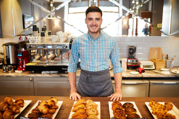 Escriturário de padaria jovem em avental de pé por balcão com grande variedade de pastelaria fresca
 - Foto, Imagem