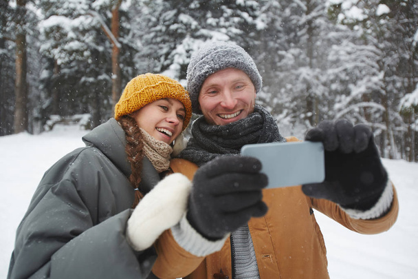 Εύθυμο ενθουσιασμένο όμορφο νεαρό ζευγάρι σε ζεστά ρούχα που στέκονται στο χειμερινό δάσος και χαμογελούν ενώ λαμβάνουν smartphone selfie - Φωτογραφία, εικόνα