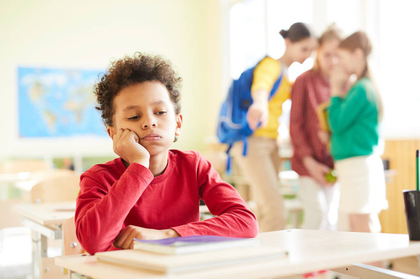 Bullying escolar: triste chico africano con el pelo rizado sentado en el escritorio y mirando hacia abajo mientras sus compañeros de clase discuten sobre él en segundo plano
 - Foto, imagen