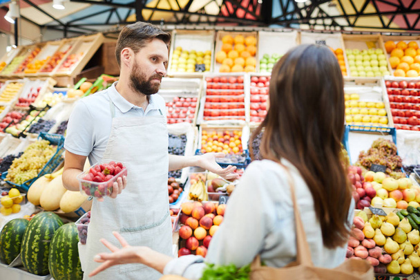 Confused misunderstanding young male grocer in apron holding container of strawberries shrugging shoulders while talking to customer in farmers market - Foto, imagen