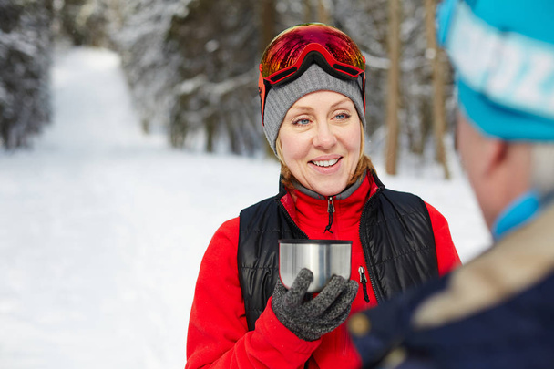 Ευτυχισμένος ώριμη γυναίκα σε activewear κρατώντας φλιτζάνι καυτό τσάι, ενώ μιλώντας για το σύζυγό της μετά το σκι κατάρτισης - Φωτογραφία, εικόνα