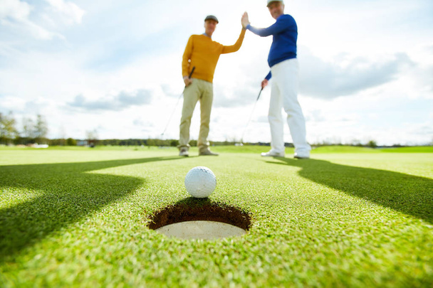 Two active men making high five while standing on play field and looking at golf ball by hole - Photo, Image