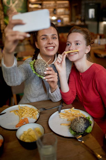 Two friendly girls making funny selfie with fast food while having lunch in cafe - Photo, Image