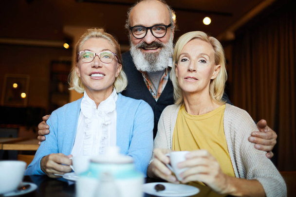 Ανώτερος γενειοφόρος άνδρας αγκαλιάζει δύο ξανθιά μεσήλικες γυναίκες κατά τη διάρκεια τους στέκι στο καφέ - Φωτογραφία, εικόνα