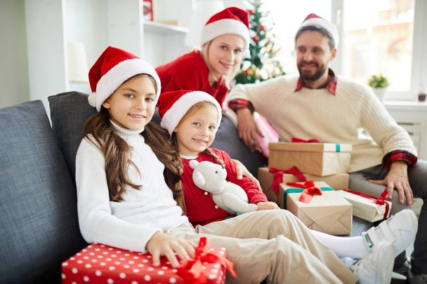 クリスマス前夜に贈り物の山とソファに座ってサンタキャップの小さな女の子とその両親 - 写真・画像