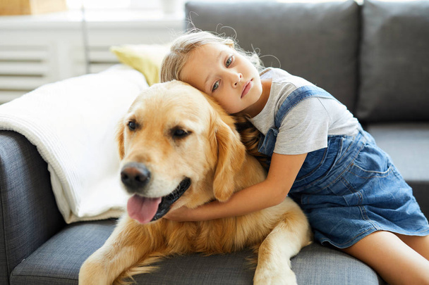 Adorable little girl embracing golden labrador while sitting next to him on sofa at home - Photo, Image