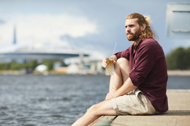 Joven hombre relajado con vaso de cóctel sentado en pontón por el agua en el día de verano
 - Foto, imagen