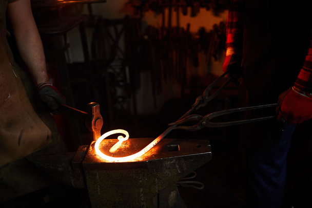 Крупный план неузнаваемых рабочих, стоящих на наковальне и делающих скрученную форму металлического стержня с помощью молотка и щипцов в мастерской
 - Фото, изображение