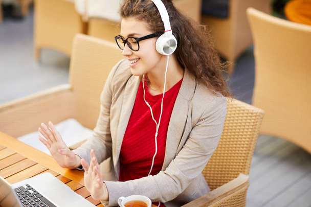 Young cheerful female in headphones talking through video-chat while sitting in cafe - 写真・画像