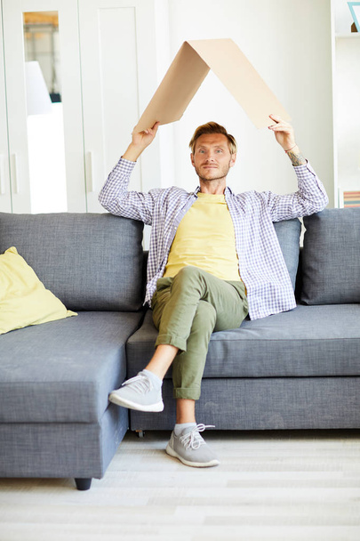 Young casual man holding carton roof over his head while sitting in living-room - Foto, Imagen
