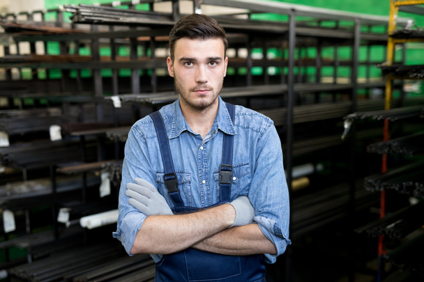 Porträt eines jungen Arbeiters, der im Overall steht, die Arme in der Stahlfabrik verschränkt - Foto, Bild