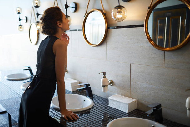 Joven hembra en vestido negro tocando su cuello mientras se mira en el espejo en el baño
 - Foto, imagen