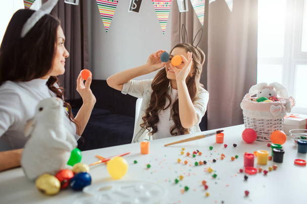 Mother and daughter prepare for Easter. Girl hold colorful eggs in front eyes. Mother does the same. They have fun. Decoration and paint on the table. - Foto, Imagem