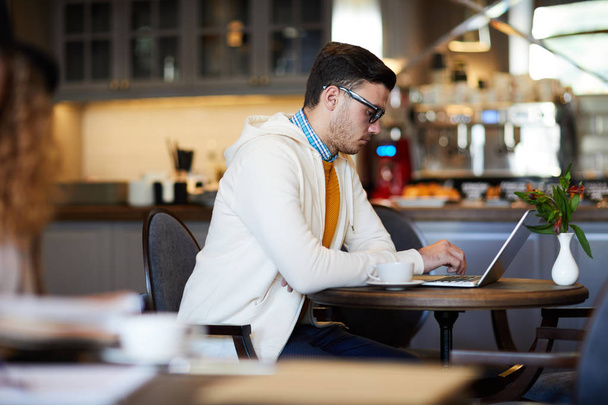 Joven emprendedor ocupado, freelancer o blogger sentado junto a la mesa en la cafetería frente a la computadora portátil y navegando por la red
 - Foto, imagen