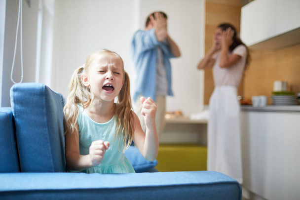 Маленька примхлива дівчинка висловлює своє незадоволення гучним голосом і вдарив крісло з шокованими батьками на фоні
 - Фото, зображення