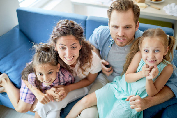 Jovens pais e meninas com expressão facial tensa sentados no sofá na frente do aparelho de tv durante a transmissão
 - Foto, Imagem