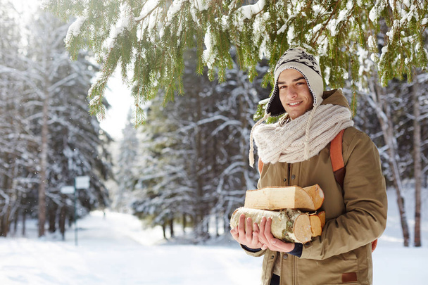 Waist up portrait of handsome young man posing in winter forest holding logs and smiling at camera cheerfully, copy space - 写真・画像