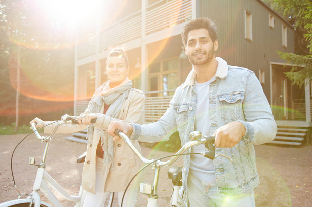 Alegre hermosa pareja joven de ciclistas en ropa casual caminando con bicicletas juntos y mirando a la cámara a la luz del sol al aire libre
 - Foto, Imagen