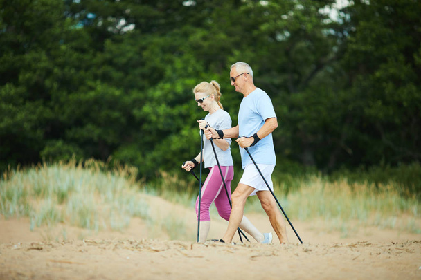 Modern Senior paar in zonnebrillen en Activewear trekking op zandstrand met groene bomen op de achtergrond - Foto, afbeelding