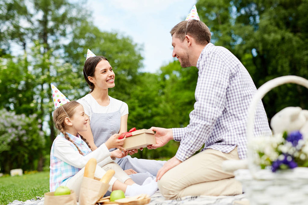 glückliches kleines Mädchen nimmt Geschenkbox aus den Händen ihres Vaters beim Geburtstagspicknick an einem Sommertag - Foto, Bild