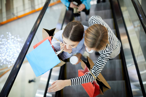 Vista superior de dos compradores felices moviéndose hacia arriba en escaleras mecánicas dentro del centro comercial moderno durante las compras
 - Foto, imagen