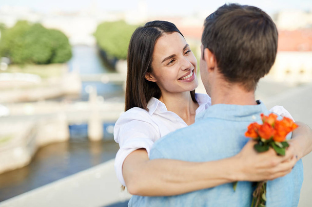 Smiling affectionate girl looking with love at boyfriend and hugging him while holding small bouquet, they having romantic date on roof - 写真・画像