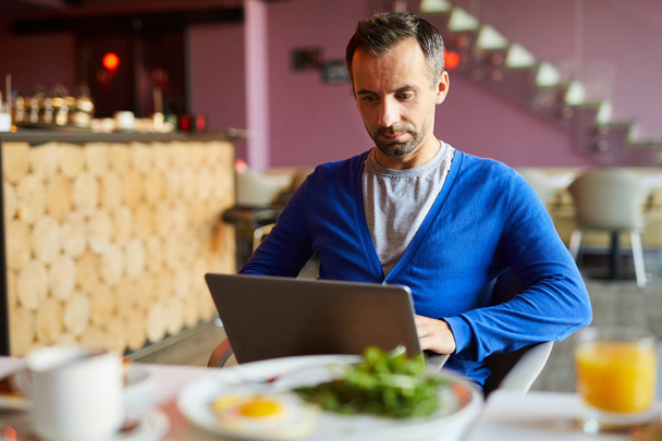 Grave guapo moderno freelancer masculino con rastrojo utilizando el ordenador portátil mientras se prepara informe en la cafetería
 - Foto, Imagen
