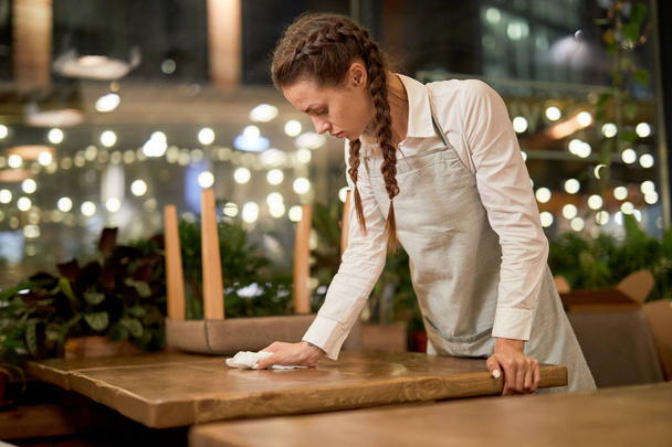 Young waitress in apron cleaning wooden table after the last clients in the evening - Фото, изображение