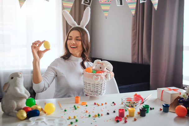 Happy positive young woman prepare fo Easter alone. She sit at table in room and hold yellor egg. Basket in front. She smile. Decoration and sweets on table. - Foto, Imagem