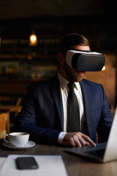 Virtuální realita: vážný koncentrovaný vousatý obchodník ve VR brýlemi pomocí dotykové podložky na laptopu při správě interaktivního zařízení v kavárně - Fotografie, Obrázek