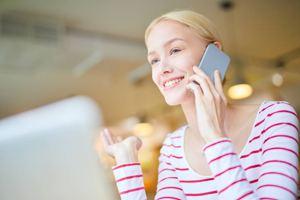 junge Frau mit zahmem Lächeln spricht per Smartphone, während sie vor dem Laptop sitzt - Foto, Bild