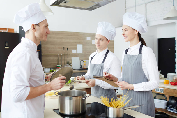 Estagiários confiantes alegres em uniformes sorrindo enquanto conversam com o chef profissional e fazem anotações na escola de culinária enquanto ouvem conselhos de chef qualificado
 - Foto, Imagem