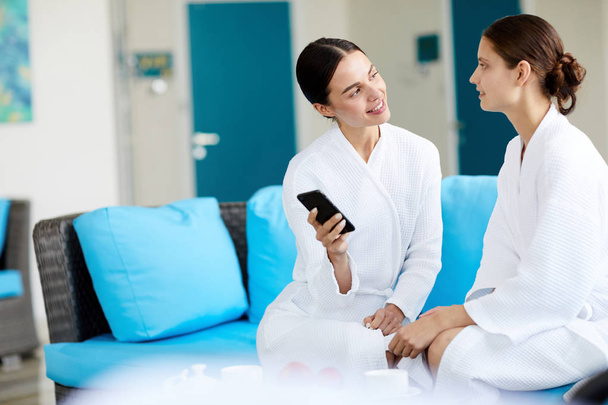 One of young women showing her friend notification in smartphone and discussing it in spa center - Foto, Bild