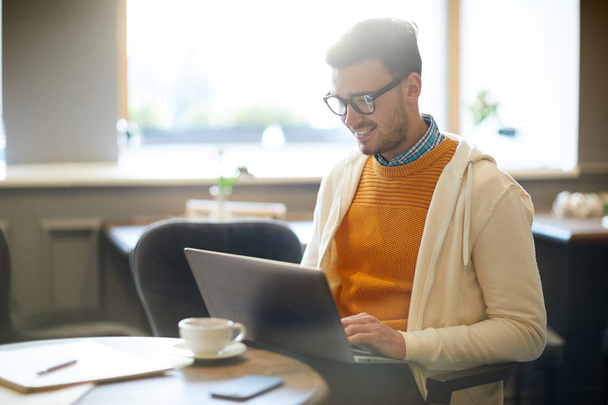Щасливий молодий блогер з ноутбуком сидить в кафе і переглядає в мережі за чашкою кави
 - Фото, зображення