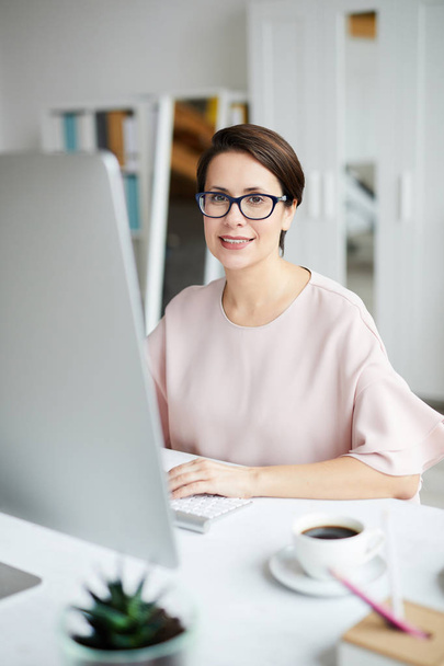 junge selbstbewusste Geschäftsfrau sitzt während der Arbeit an ihrem Arbeitsplatz vor dem Monitor - Foto, Bild