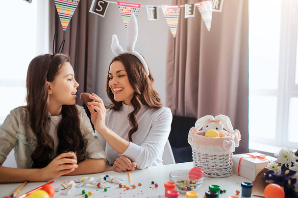 Mother and daughter prepare for Easter. They sit at table in room. Young woman feed girl with chocolate egg. Kid keep mouth opened. Decoration and paint with sweets on table. - Photo, Image