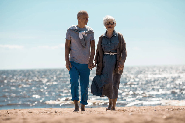 Босоногі літні люди, що йдуть піщаним пляжем влітку, охолоджуються в сонячний день на курорті
 - Фото, зображення