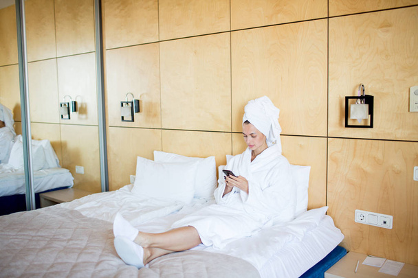 Fiatal pihentető nő, fehér fürdőköpeny, törölköző a fej és a békaláb ül az ágyban, és textil a smartphone - Fotó, kép