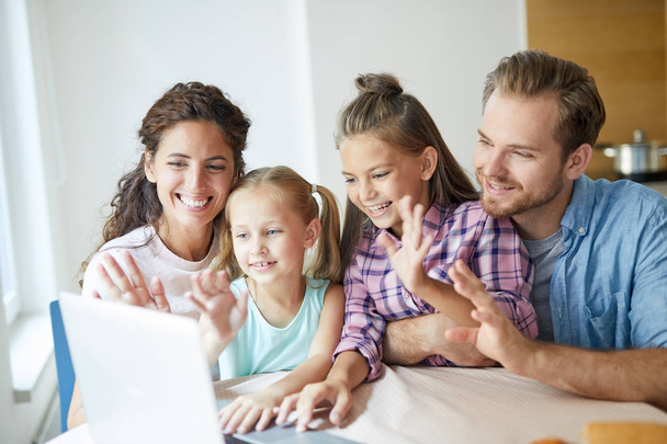 Jovem família alegre moderna de quatro pessoas sentadas à mesa na frente do laptop e conversando por vídeo-chat com seus amigos ou parentes
 - Foto, Imagem