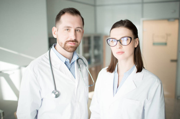 Twee jonge clinici in whitecoats kijken naar camera in hedendaagse klinieken - Foto, afbeelding
