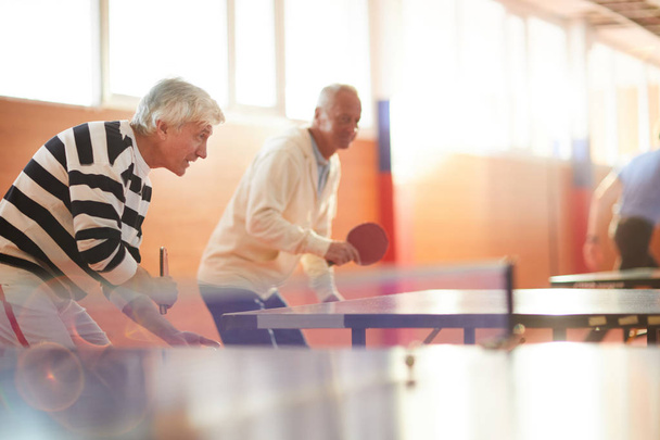 Dois homens ativos seniores com raquetes tentando pegar bola de ping pong durante o jogo de lazer
 - Foto, Imagem