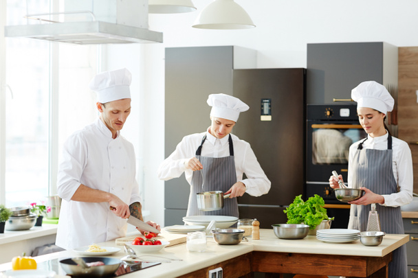 Chef ocupado e seus cozinheiros que trabalham na cozinha e cozinhar macarrão no balcão: chef de corte de tomates, jovem em avental verificando espaguete, sério senhora chicotadas ingredientes
 - Foto, Imagem