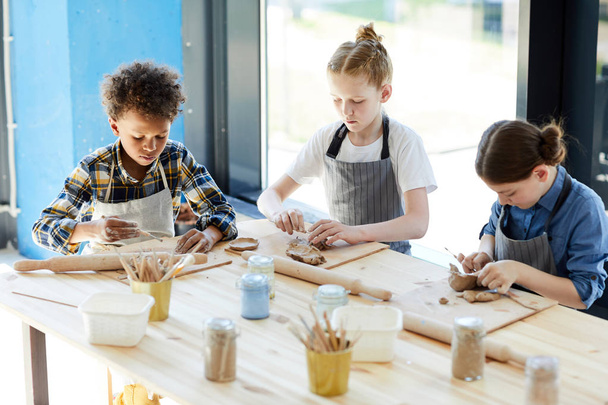 Tre bambini in grembiuli seduti a tavola, impastando argilla e facendo terracotta a lezione
 - Foto, immagini