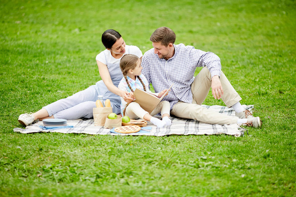Молоді батьки та їх маленька донька сидять на зеленому газоні, розслабляючи та читаючи книгу казок під час пікніка
 - Фото, зображення