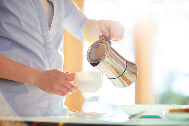 Mulher anônima derramando água quente do pote na xícara ao fazer café para o café da manhã na cozinha iluminada pelo sol
 - Foto, Imagem