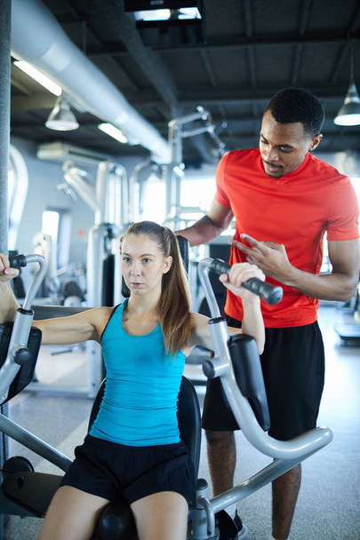 Young active woman exercising on sports equipment in gym with her trainer standing near by - Photo, Image