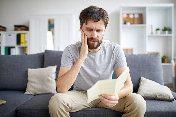 Embarrassé homme lecture papier ou facture de paiement et toucher son visage tout en étant assis sur le canapé à la maison
 - Photo, image