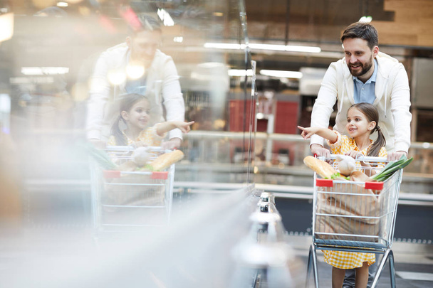 Menina positiva apontando para o produto na prateleira enquanto empurra carrinho de compras junto com o pai bonito no supermercado
 - Foto, Imagem