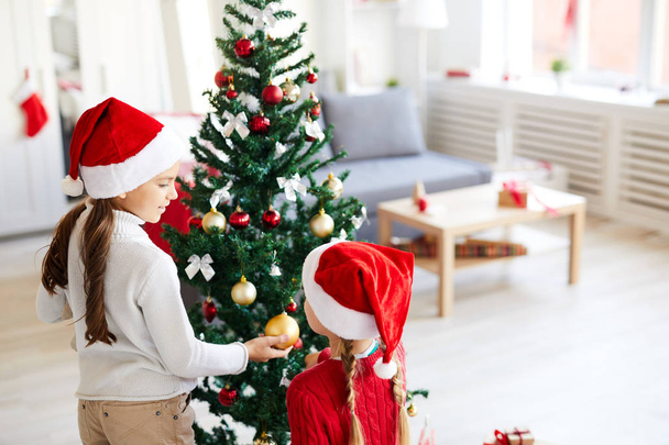 eines der süßen Weihnachtsmädchen zeigt ihrem Geschwister goldene Kugel auf geschmücktem Tannenzweig - Foto, Bild